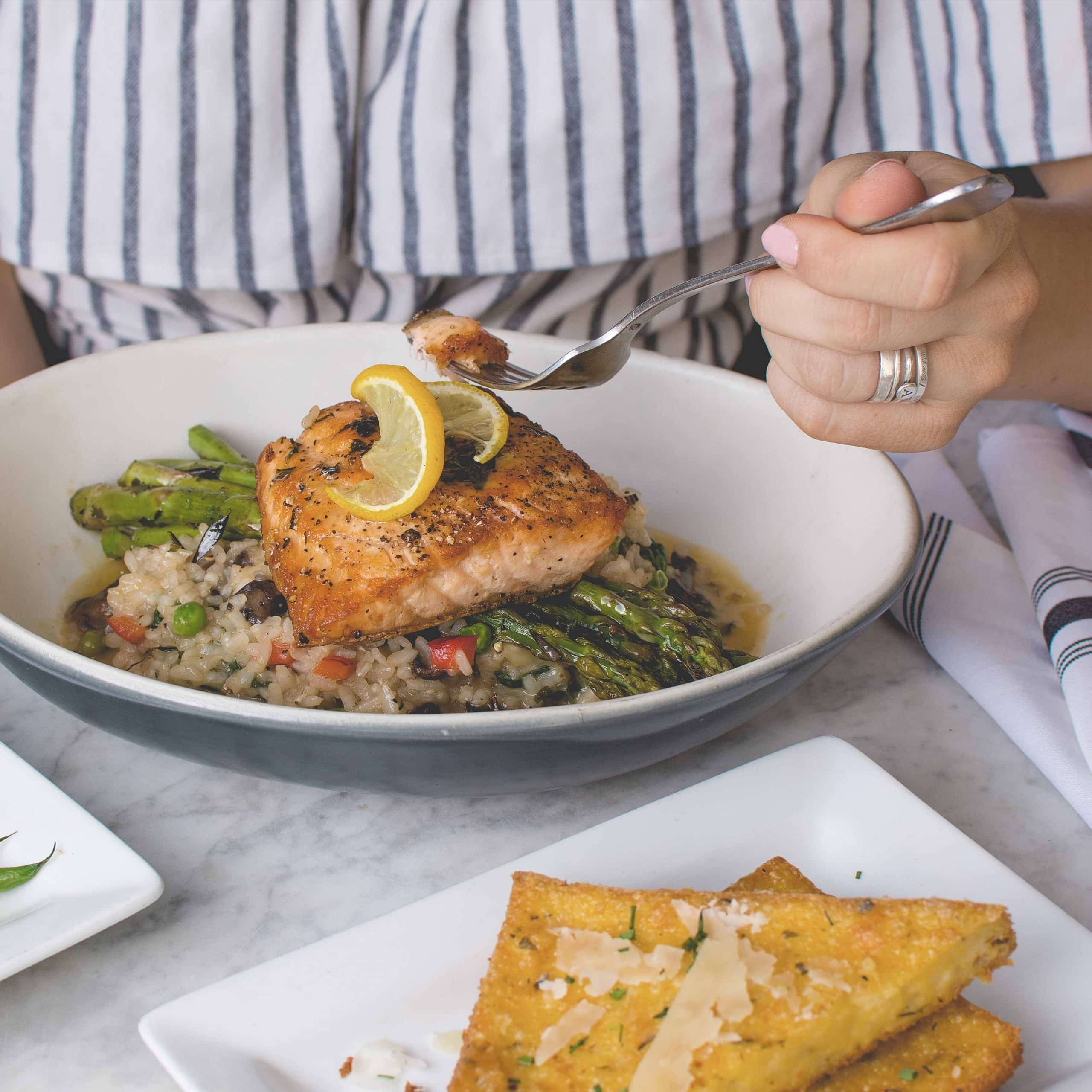 Plato con una base de arroz y verduras con un salmón encima mientras una mano femenina sostiene un tenedor al lado